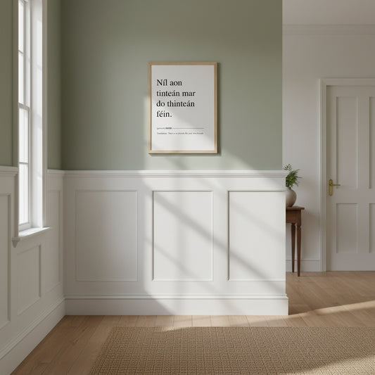 Níl aon tinteán mar do thinteán féin. Translation: There Is No Fireside Like Your Own Fireside. This Irish language framed print makes a great present, birthday gift and ideal Irish housewarming gift, or for Gaelic speakers. Gaelic proverb art ‘Níl aon tinteán mar do thinteán féin’ in natural oak frame, cozy hallway with wainscoting, Farrow & Ball-style muted green paint, woven runner, warm morning light, lifestyle product shot.