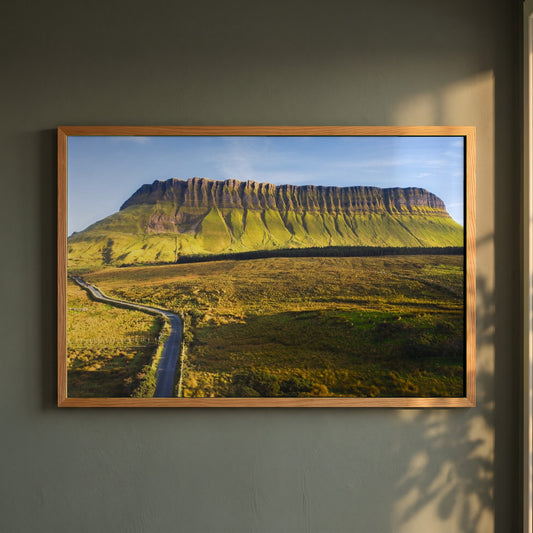 Benbulben, Sligo framed print in a natural oak frame on a dark green wall beside a window, dappled plant shadow