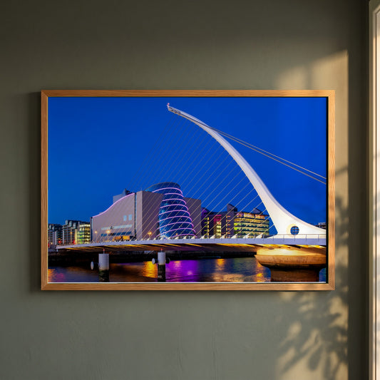 Samuel Beckett Bridge Photography framed print in a natural oak frame on a dark green wall beside a window, dappled plant shadow