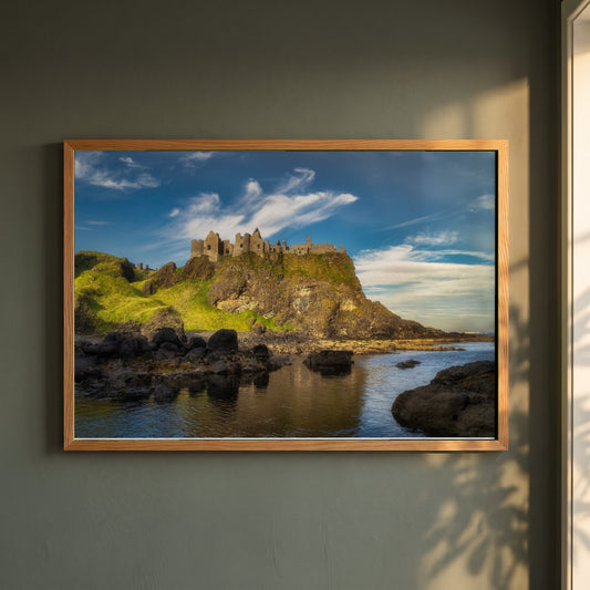 Dunluce Castle framed print in a natural oak frame on a dark green wall beside a window, dappled plant shadow