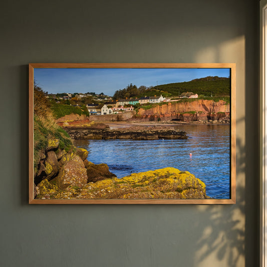 Dunmore East framed print in a natural oak frame on a dark green wall beside a window, dappled plant shadow