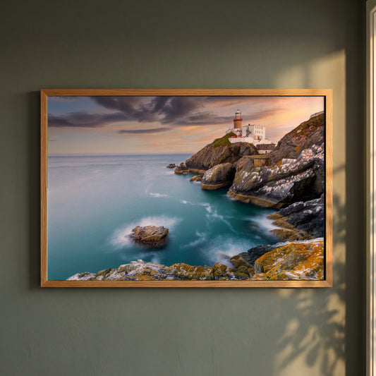 Baily Lighthouse Sunset, Howth Head framed print in a natural oak frame on a dark green wall beside a window, dappled plant shadow