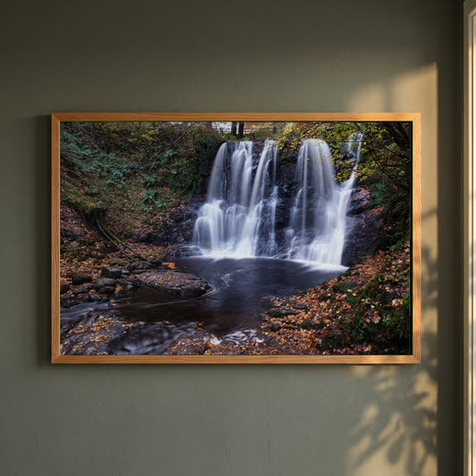 Glenariff Waterfall framed print in a natural oak frame on a dark green wall beside a window, dappled plant shadow