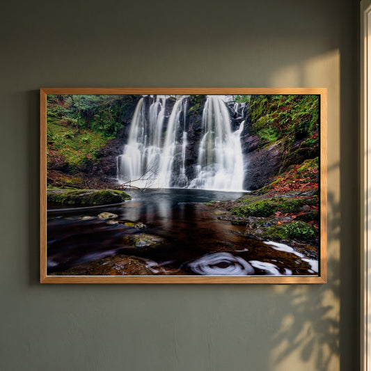 Glenariff Waterfall framed print in a natural oak frame on a dark green wall beside a window, dappled plant shadow