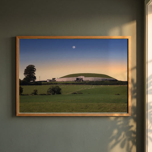 Newgrange Irish Sunset framed print in a natural oak frame on a dark green wall beside a window, dappled plant shadow