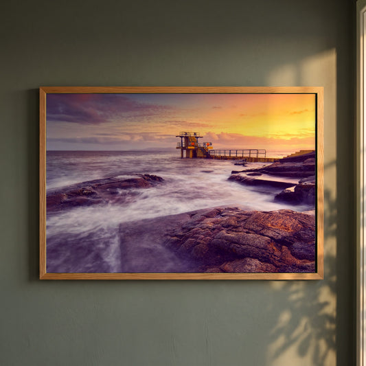 Salthill Diving Tower Galway framed print in a natural oak frame on a dark green wall beside a window, dappled plant shadow