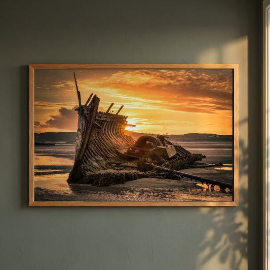 Sunset Ship Wreck Bád Eddie Bunbeg framed print in a natural oak frame on a dark green wall beside a window, dappled plant shadow