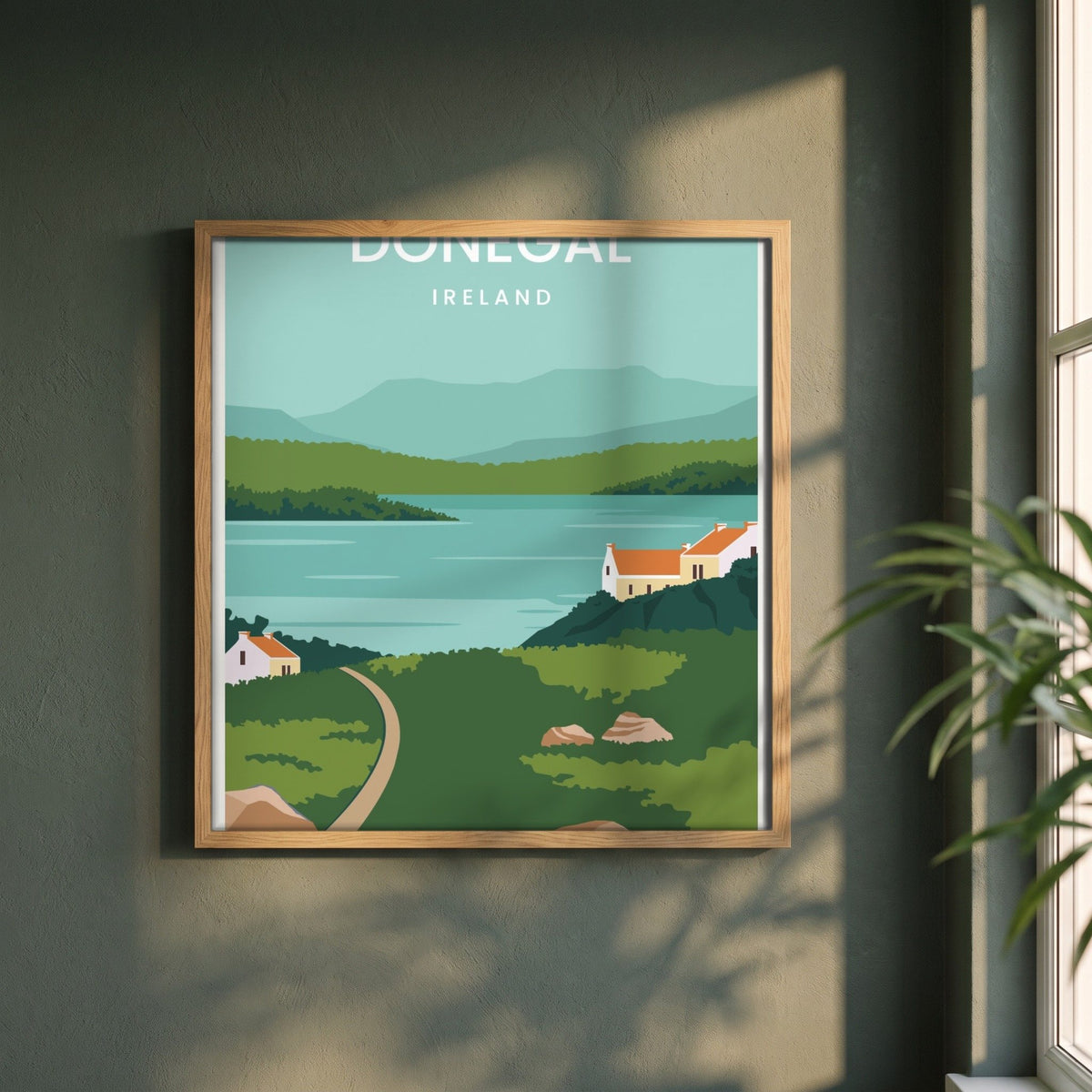 Donegal minimalist travel print in a natural oak frame on a dark green wall beside a window, dappled plant shadow