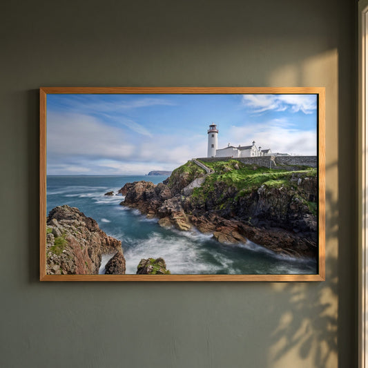 Fanad Head Lighthouse, Co. Donegal framed print in a natural oak frame on a dark green wall beside a window, dappled plant shadow