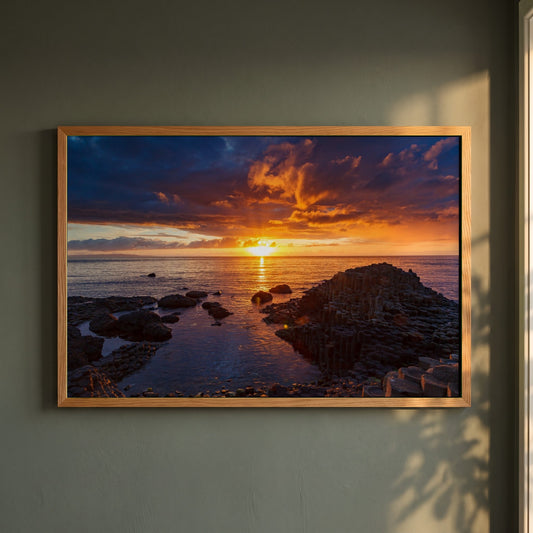 Giant's Causeway framed print in a natural oak frame on a dark green wall beside a window, dappled plant shadow