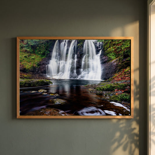 Glenariff Waterfall framed print in a natural oak frame on a dark green wall beside a window, dappled plant shadow