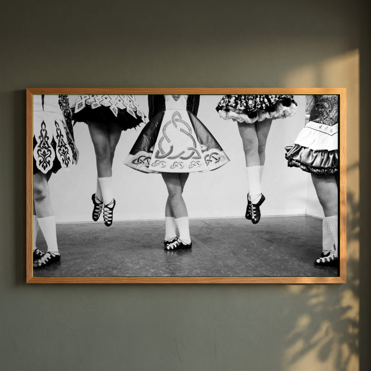 Irish Dancing Black & White framed print in a natural oak frame on a dark green wall beside a window, dappled plant shadow