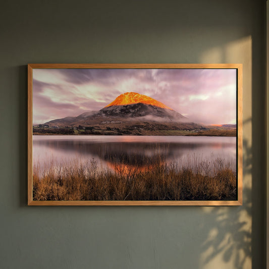 Orange Glow on Mount Errigal at Sunset framed print in a natural oak frame on a dark green wall beside a window, dappled plant shadow