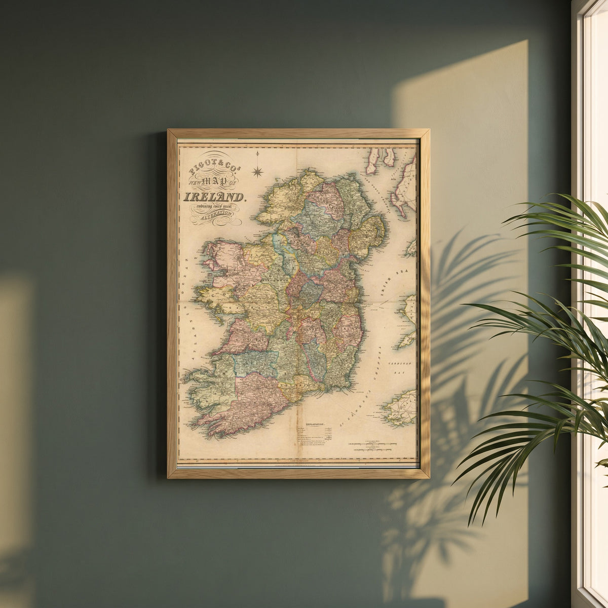 Map of Ireland framed print in a natural oak frame on a dark green wall beside a window, dappled plant shadow