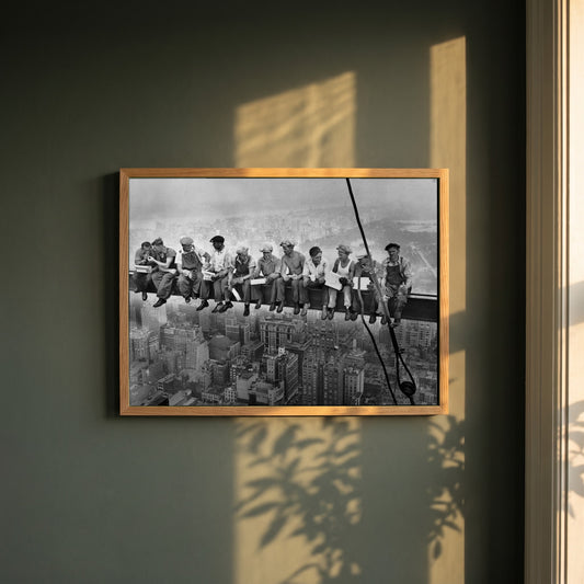 Lunch Atop a Skyscraper framed print in a natural oak frame on a dark green wall beside a window, dappled plant shadow