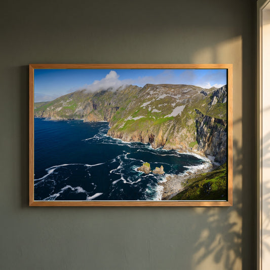 Slieve League framed print in a natural oak frame on a dark green wall beside a window, dappled plant shadow