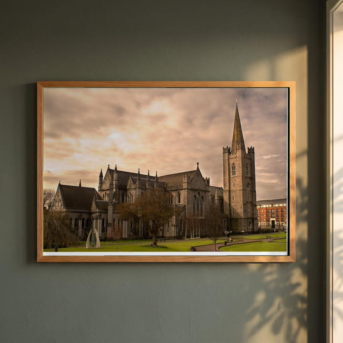 St. Patrick's Cathedral Dublin framed print in a natural oak frame on a dark green wall beside a window, dappled plant shadow